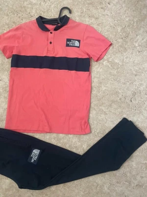 Rosa och marinblå polotopp från The North Face - Säljer en snygg polotopp från The North Face i rosa med en bred marinblå rand över bröstet och matchande marinblå krage. Toppen har korta ärmar, knappar framtill och en tydlig logga på bröstet. Perfekt för dig som gillar sportig och stilren design.