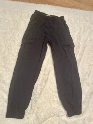 Mörkblå cargopants från Lindex, 116 - Mörkblå cargopants från Lindex med elastisk midja och muddar vid benslut. Byxorna har praktiska sidofickor och cargofickor på benen. Tillverkade i mjuk bomull som är skön att ha på sig hela dagen. Perfekt för dig som gillar en avslappnad och cool stil.