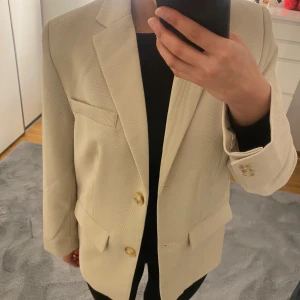 Beige kavaj från NA-KD - Snygg beige kavaj från NA-KD med klassisk krage och två guldiga knappar framtill. Kavajen har fickor med lock, slits bak och är helfodrad. Passformen är loose och den har långärmade ärmar. Perfekt för dig som gillar stilrena och trendiga plagg.