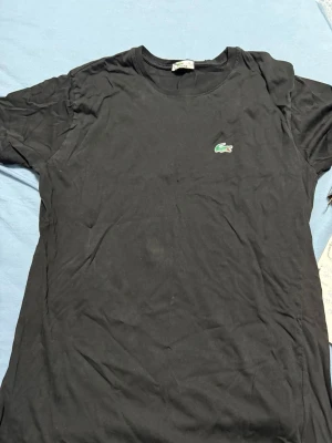 Svart t-shirt från Lacoste, storlek L - Svart t-shirt från Lacoste med klassisk rund hals och korta ärmar. På bröstet sitter den ikoniska gröna krokodilloggan. T-shirten är gjord i mjuk bomull och har en clean, enkel look som funkar till allt. Perfekt för dig som gillar stilrena plagg med diskret branding.