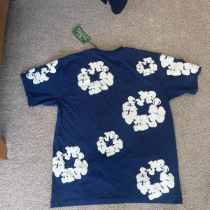 Blå t-shirt med vita blommor - Snygg blå t-shirt från Denim tears med stora vita blommiga tryck över hela tröjan. Klassisk passform och korta ärmar, perfekt för dig som vill sticka ut med en cool och unik stil.