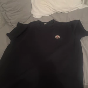 Svart Moncler t-shirt med logga - Svart t-shirt från Moncler med klassisk rund hals och korta ärmar. På bröstet sitter Monclers ikoniska logga i vitt och rött. T-shirten är gjord i mjuk bomull och har en clean, stilren look som funkar till allt. Perfekt för dig som gillar exklusiva märken.