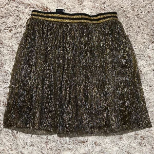 Glittrig kjol från H&M Divided - kjol med glittrigt, metalliskt tyg i guld och svart från H&M Divided. Kjolen har resår i midjan med svarta och guldiga ränder, samt ett tunt yttertyg med diskret ränder. Fodrad insida i svart polyester för extra komfort.  Kjolen har två lager (se bild 3) Storlek M
