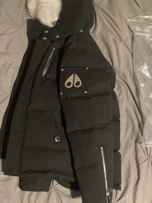 Svart pufferjacka Moose Knuckles - Säljer en svart pufferjacka från Moose Knuckles med stor vit pälskant på huvan och ikonisk logga på ärmen. Jackan har flera dragkedjor, knappar och fickor samt en schysst oversized look. Perfekt för kalla dagar och riktigt snygg streetstil.