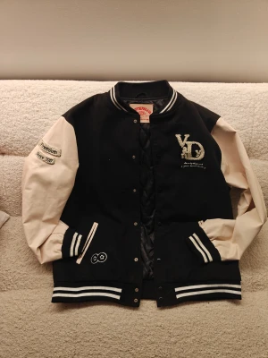 Svart och beige bomberjacka VandyThePink - Säljer en riktigt snygg svart och beige bomberjacka från VandyThePink & Co. Jackan har broderade detaljer, blommor på ryggen och coola patches på ärmarna. Ribbstickade muddar och krage med vita ränder. Materialmix av bomull och polyester. Perfekt för dig som vill sticka ut.