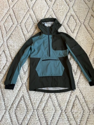 Skidjacka från Five Seasons, blå/svart - Snygg skidjacka från Five Seasons i blått och svart med huva och half zip. Jackan har stor ficka framtill med dragkedja och extra dragkedja på ärmen och på andra ställen. Materialet är syntet och jackan är både vattentät och vindtät, perfekt för blåsiga dagar. Är använd fåtalsgånger.