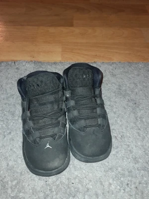 Svarta Jordan sneakers för barn - Säljer ett par svarta Jordan sneakers med högt skaft och snörning. Skorna har en rund tå, platt sula och är tillverkade i en kombination av mocka och syntetmaterial. Jumpman-loggan syns på tån och hälen. Perfekt för dig som gillar streetstyle och sportig look.