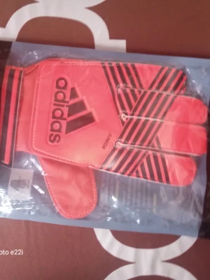 Adidas ACE Junior målvaktshandskar - Säljer ett par Adidas ACE Junior målvaktshandskar i en snygg röd färg med svarta ränder och Adidas-logga. Handskarna har justerbar kardborre vid handleden och är designade för bra grepp och komfort vid fotbollsspel. Perfekt för unga målvakter som vill ha stil och funktion.