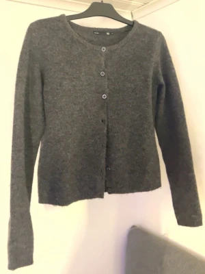 Mörkgrå stickad kofta - Säljer en mörkgrå stickad kofta från lager 157 i storlek S. Koftan har rund hals, långa ärmar och knäppning med fem knappar framtill. Materialet är mjukt och värmande, perfekt för kyliga dagar. Enkel och stilren design som funkar till många olika outfits.