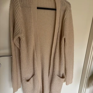 Beige stickad kofta från Gina Tricot - Mysig beige stickad kofta från Gina Tricot i storlek XS. Lång modell med två stora fickor framtill och ribbade muddar. Perfekt att slänga på sig över en t-shirt eller topp när det är kyligt ute.