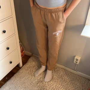 Beige mjukisbyxor från 157, S - Beiga mjukisbyxor från lager 157 med tryck 'South Beach Padel Sports Club' i vitt och ljusblått på ena benet. Byxorna har resår i midjan och vid bensluten, samt mjuk fleece på insidan för extra komfort. 