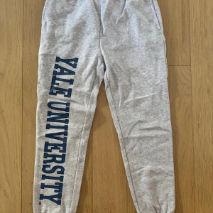 Grå Yale University Sweatpants H&M - Grå sweatpants från H&M x Yale University med tryck längs ena benet. Byxorna har resår i midjan och är tillverkade i en mjuk bomulls- och polyestermix.
