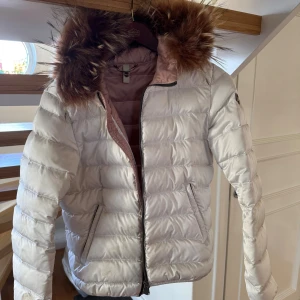 Vit pufferjacka med pälskrage från Colmar - Säljer en snygg vit pufferjacka från Colmar med ljusrosa foder och stor huva med pälskrage. Jackan har dragkedja framtill, två fickor med dragkedja och quiltad design. Perfekt för kalla dagar och riktigt mysig att bära. Jackan har tyvärr fått en reva i tyget på ena armen, se bild. Detta går att lämna in och fixa snyggare. Storlek på jackan passar S/ liten M