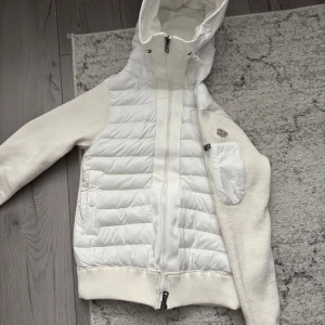 Vit cardigan från Moncler med huva - Säljer en snygg vit cardigan från Moncler i storlek S (helt ny). Jackan har stickade ärmar, quiltad front, dragkedja framtill och en praktisk huva. Moncler-logga på ärmen (Nfc-scan funkar) och cool detalj med ficka på sidan. Perfekt för kyliga dagar när du vill vara både varm och stilren.