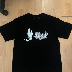 Svart t-shirt från VLONE med tryck - Svart t-shirt från VLONE i bomull med stor vit V-tryck på ryggen och coolt tryck med fe och text på bröstet. Klassisk passform och korta ärmar. Perfekt för dig som gillar streetwear och vill sticka ut med en unik design.
