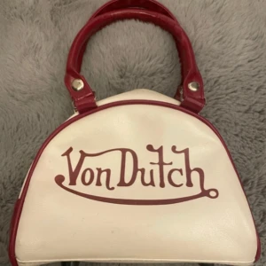 90s-00s Von Dutch handväska - Cool handväska från Von Dutch i vit och röd färg med klassisk logga framtill. Väskan har rundad form, röda handtag och detaljer samt dragkedja upptill. Tillverkad i syntetmaterial med retrovibbar, perfekt för dig som gillar 2000-tals mode! 