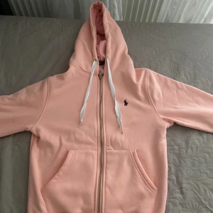 Rosa hoodie från Polo Ralph Lauren - Mysig ljusrosa hoodie från Polo Ralph Lauren med dragkedja, vit snörning i huvan och klassisk logga broderad på bröstet. Tröjan har känguruficka och är fodrad med mjuk fleece på insidan. Perfekt för chill dagar.
