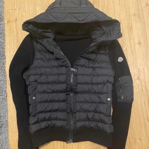 Svart Moncler cardigan med huva - Svart cardigan från Moncler med stickade ärmar och quiltad kropp. Jackan har huva, dragkedja framtill, fickor med tryckknappar och en extra ficka med dragkedja på ärmen. Klassisk Moncler-logga på vänster ärm. Perfekt för kyliga dagar.