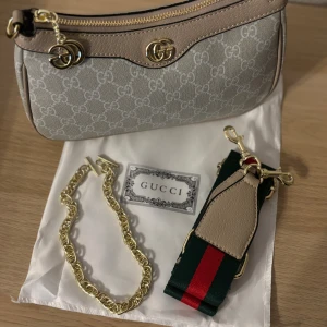 Gucci beige axelväska med GG-mönster - Snygg beige axelväska från Gucci med klassiskt GG-mönster i vitt och detaljer i ljusbrunt skinn. Väskan har guldfärgade metalldetaljer, dragkedja och medföljer både en chunky guldkedja och en bred axelrem i grönt och rött. Perfekt för dig som vill ha en ikonisk och trendig accessoar.