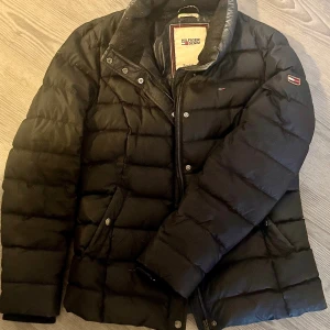Svart pufferjacka från Hilfiger Denim - Svart pufferjacka från Hilfiger Denim med klassisk quiltad design och diskreta logodetaljer på bröstet och ärmen. Jackan har dragkedja och tryckknappar framtill samt två fickor. Perfekt för kalla dagar och riktigt snygg till streetwear.