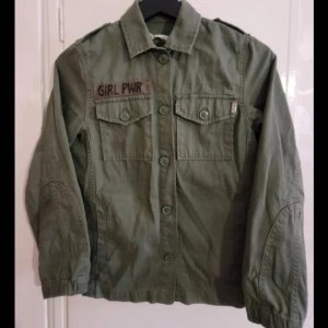 Grön overshirt från H&M med GIRL PWR - Stilren militärgrön overshirt från H&M med coolt 'GIRL PWR'-tryck på bröstet och peace-märke på ärmen. Jackan har klassisk krage, knäppning framtill, två bröstfickor med lock och långa ärmar med resår vid ärmslut. Perfekt för en avslappnad streetstyle.