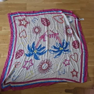 Färgglad sjal från Liu Jo Milano - Stor fyrkantig halsduk från Liu Jo Milano med lekfulla motiv som solar, hjärtan, stjärnor och blommor. Basen är vit med detaljer i rosa, blått, rött och gult samt en bred rosa kant. Tillverkad i silke, perfekt att knyta runt halsen eller håret för en cool look.