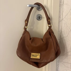 Brun Marc Jacobs väska -  En handväska i fint skick från Marc Jacobs. Den är perfekt att ha över axeln eller som handväska. Väskan har guldiga metalldetaljer, mjuk form och ett stort innerfack.  Bredd:28cm  Höjd:23cm  Djup::10cm  Måtten är inte exakta  Skriv vid fler funderingar💗