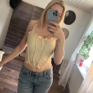 Beige spetsig korsett-topp med rosett - Säljer en beige korsett-topp i spets med smala axelband och söt rosettdetalj framtill. Toppen är croppad och har en feminin, figurnära passform med dekorativa sömmar som ger en snygg siluett. Perfekt för dig som gillar romantisk och trendig stil. Mjukt material.