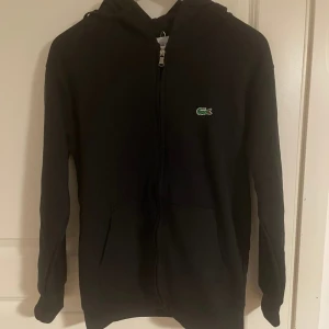 Svart hoodie från Lacoste med dragkedja - Svart hoodie från Lacoste med klassisk krokodillogga på bröstet. Tröjan har huva, hel dragkedja framtill och två fickor. Perfekt för en chill och stilren look. Tillverkad i mjuk bomull och har långa ärmar.