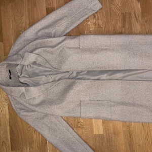 Ljusgrå kappa med bälte från Gina Tricot - Stilren ljusgrå kappa från Gina Tricot med klassisk krage, raka linjer och två stora fickor framtill. Kappan har ett matchande bälte i midjan och är helfodrad för extra komfort. Perfekt för dig som gillar minimalistisk och clean stil.