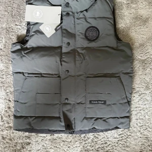 Canada Goose Väst - Exklusiv väst från Canada Goose Black Label i elegant grå färg. En lyxig och funktionell premiumväst som kombinerar värme och stil – perfekt för både vardag och fritid.  ♦️ Helt ny – med originaltag kvar ☑️ ⚜️ Storlek M – normal passform ☑️ ♦️ Black Label – exklusiv och stilren design ☑️ ⚜️ Praktiska fickor & tidlös look ☑️  📦 Snabb leverans & trygg affär 