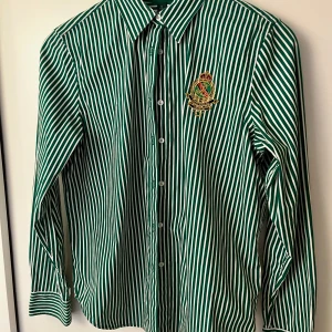 Grönvit randig skjorta Ralph Lauren - Snygg grön och vit randig skjorta från Ralph Lauren med broderad crest på bröstet. Skjortan har klassisk krage, knäppning framtill och långa ärmar med manschett. Tillverkad i 100% bomull för en skön känsla.