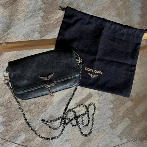 Svart axelväska från Zadig & Voltaire - Snygg svart axelväska i skinn från Zadig & Voltaire med silverfärgad kedjerem och ikoniskt vingemblem framtill. Tyvärr har den lilla silver kedjan gått av. Väskan har dragkedja både fram och bak samt detaljer i silver. Perfekt storlek för det viktigaste och ger en cool vibe till din outfit.