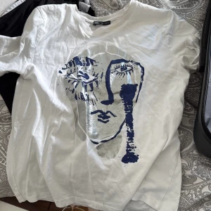 Vit t-shirt med konstnärligt tryck från Zara - Vit t-shirt från Zara med ett stort konstnärligt ansikte i silver och mörkblått framtill. Klassisk rund hals och korta ärmar. Skön passform i mjuk bomull, perfekt för dig som gillar unika prints och stilrena vibes.