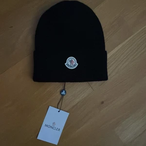 Svart Moncler mössa i ull - Svart ribbstickad mössa från Moncler med klassisk uppvikt kant och broderad logotyp framtill. Tillverkad i mjuk ull som håller dig varm under kyliga dagar. Perfekt accessoar för en clean och stilren look.