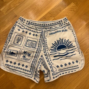 Sommar Club vita mönstrade shorts - Säljer ett par vita shorts från Sommar Club med blått grafiskt mönster och tryck, bland annat sol, palmer och blommor. De har elastisk midja och är gjorda i 100% polyester, perfekta för strand eller chill dagar. Snygg och unik design som sticker ut.