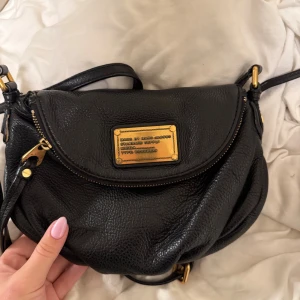 Svart axelväska från Marc Jacobs - Snygg svart axelväska från Marc Jacobs i äkta skinn med guldfärgade metalldetaljer och dragkedja. Väskan har en rundad form och ett stort fack med coolt mönstrat innerfoder med bokstäver. Perfekt storlek för det viktigaste och har justerbar axelrem.