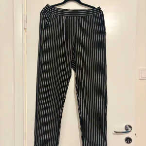 Svarta randiga byxor med vit pinstripe - Svarta byxor med smala vita vertikala ränder och bred resår i midjan. Modellen är loose fit med raka ben och uppvikta benslut. Snyggt och stilrent mönster som passar till många olika looks. Tillverkade i ett mjukt och lätt material. Storlek 170 men passar även XS