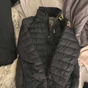 Svart lättviktsjacka Parajumpers YS - Svart quiltad lättviktsjacka från Parajumpers med gul detalj vid kragen och broderad patch på ärmen. Jackan har dragkedja framtill, ståkrage och en bröstficka med dragkedja. Perfekt för dig som vill ha en snygg och funktionell jacka.