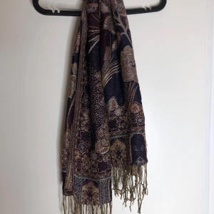 Blommig mörkblå halsduk med fransar - Snygg halsduk i mörkblått med detaljerat blommönster i beige och bruna toner. Halsduken har långa fransar i ändarna och ett elegant, bohemiskt uttryck. Perfekt accessoar för att lyfta din outfit med lite extra stil.