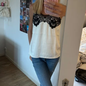 Vit linnetopp med svart spets - Säljer en vit linnetopp med snygg svart spets upptill. Toppen har en rak passform och är ärmlös med tunna axelband. Materialet är glansigt och känns lyxigt, perfekt för att lyfta vilken outfit som helst. Spetsen har ett detaljerat blommönster som ger en extra touch.