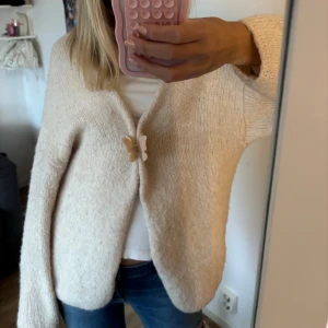 Beige stickad kofta med fjärilsspänne - Supermysig beige stickad kofta med lång ärm och oversized passform. Koftan stängs framtill med ett gulligt fjärilsspänne som ger en söt detalj. Perfekt att slänga över en t-shirt och jeans för en chill look.