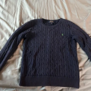 Mörkblå kabelstickad tröja Polo Ralph Lauren - Kabelstickad mörkblå tröja från Polo Ralph Lauren med rund halsringning och klassisk grön broderad logga på bröstet. Långärmad modell i bomull med ribbade muddar och nederkant. Perfekt för lager-på-lager under höst och vinter. Står L men passar även M