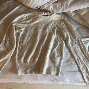 Beige stickad tröja med rosett i nacken - Supersöt beige stickad tröja med lång ärm och snygg rosettdetalj i nacken. Tröjan har ribbade muddar och en klassisk rund halsringning. Perfekt för dig som gillar stilrena och feminina detaljer.