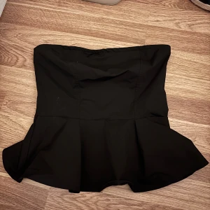 Svart peplum tubtopp - Snygg svart tubtopp med peplumvolang nertill. Toppen är axelbandslös och har en figurnära passform upptill som sedan släpper ut i en volang. Perfekt för dig som vill ha en stilren och trendig look.