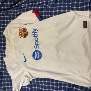 Vit FC Barcelona Nike fotbollströja - Säljer en vit FC Barcelona fotbollströja från Nike med klubbmärke, blå Nike-logga och Spotify som sponsor på bröstet. Tröjan har korta ärmar och detaljer i rött och blått på ärmslutet. Materialet är lätt och andas, perfekt för match eller träning.