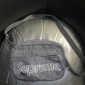 Snygg svart supreme väska - Supreme axelväska i svart med quiltad design och stort vitt Supreme-tryck på framsidan. Väskan har justerbar axelrem, dragkedja och praktiskt ytterfack. Köpte för ett år sen och inte använt så mycket,   skick 8/10