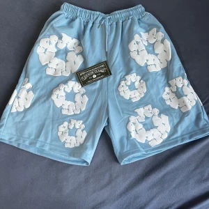 Blå shorts från Denim Tears med tryck - Säljer ett par ljusblå shorts från Denim Tears med unikt vitt tryck över hela plagget. Shortsen har elastisk midja med snörning och en bakficka. Perfekta för sommaren och riktigt sköna att bära.