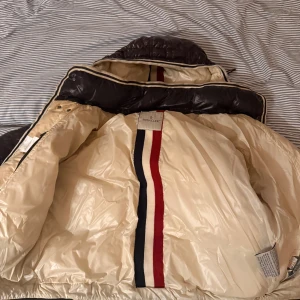 Mörkbrun dunjacka från Moncler - Säljer en mörkbrun dunjacka från Moncler med glansig finish, avtagbar huva och knappar framtill. Insidan är beige med Monclers klassiska röd, vit och blå rand. Jackan har flera fickor med dragkedja och en bröstficka med Moncler-logga.