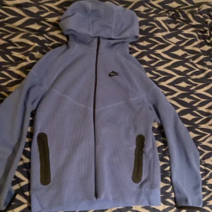Blå Nike tech fleece XS - Nike hoodiejacka i blått med svart logga på bröstet. Jackan har hel dragkedja framtill, två svarta fickor med dragkedja och huva. Materialet är syntet med rutigt mönster och känns sportigt och lätt. Perfekt för dig som gillar en clean och sportig stil. Tröjan är aldrig använd utan bara tvättad en gång. Den är storlek XL 158-170 i barn men passar XS.